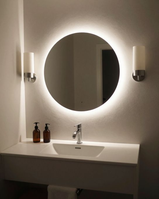 Illustration: a bathroom vanity lit by a backlit round mirror and two wall sconces at face height, evening mood